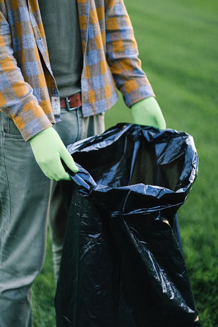 A person standing outdoors on a grassy area is holding open a large black rubbish bag lined with a shiny, metallic inner layer. The individual is wearing light green gloves, a green shirt, and a checkered jacket with shades of yellow, gray, and blue, along with khaki trousers and a brown belt. The rubbish bag appears partially filled and is positioned in front of the person's waist, with their hands gripping the sides of the bag's opening, suggesting they are in the process of collecting waste. The background features a blurred green lawn, indicating an outdoor setting suitable for waste collection or disposal, which may be part of a private rubbish removal or on-site clearance service. The scene is well-lit with natural daylight, emphasizing the textures of the bag, gloves, and clothing. This setup aligns with independent rubbish disposal practices offered by waste removal specialists such as Waste Removal Clapham, focusing on environmentally responsible waste handling and collection outside of local authority services.