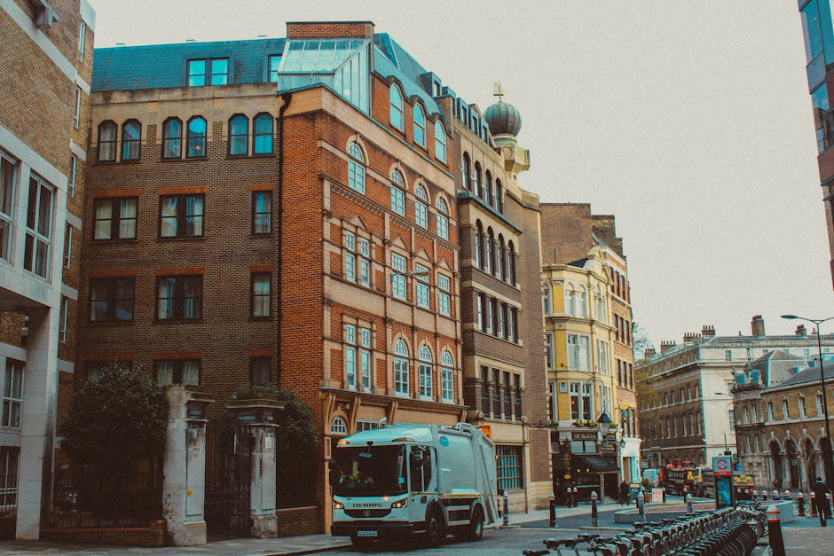 A city street scene featuring a white waste collection truck with orange and black accents parked on the paved road near a corner. The truck is positioned in front of several multi-storey buildings with varying architectural styles, including red brick facades and decorative window frames. The background includes a mix of historic and modern structures, with some buildings having rounded bay windows and ornate details, and others with flat, glass-fronted exteriors. The street is lined with black bollards and a row of bicycle racks in the foreground, and there are street lamps and signs visible along the sidewalk. The overall scene suggests an urban environment where private waste collection services like Waste Removal Clapham facilitate rubbish disposal, contrasting with council-run collections by highlighting independent waste handling activities. The lighting is natural, casting soft shadows and emphasizing the textures of the buildings' materials and the clean, organized appearance of the street.