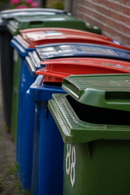 A row of large, wheelie plastic rubbish bins lined up outdoors along a paved surface next to a brick wall. The bins are predominantly blue with green, red, and dark green lids, all featuring ridged textures on top. Some bins have visible white markings or symbols on their sides. The scene depicts an arrangement typical of private waste collection services, with the bins positioned for easy access. The background shows a blurred view of a grassy area and a tree, suggesting an outdoor residential environment. Natural daylight illuminates the scene, casting soft shadows and highlighting the different colors and textures of the bins, which are indicative of different waste categories or disposal purposes typically associated with independent rubbish collection services in the local area. The image emphasizes the organized grouping of waste containers used in alternative waste handling arrangements, often seen outside homes for scheduled rubbish removal by services like Waste Removal Clapham.