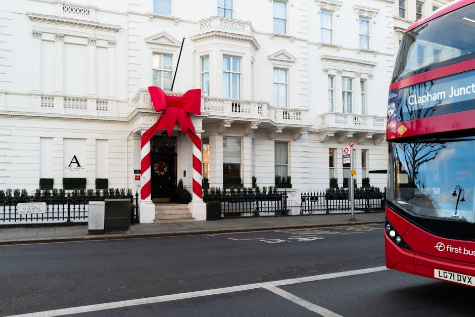 The image depicts the exterior of a white, multi-story Victorian-style building with ornate architectural details including decorative window trims, balustrades, and a small balcony. The building is decorated for the holiday season with a large red bow and candy cane-striped posts framing the entrance door, which is topped with a wreath. Small potted plants line the black iron fence that surrounds the front entrance area. The scene is set on a paved urban street with a double yellow line running along the curb, and a modern red double-decker bus is visible on the right side of the image, partially showing the front and side, with the words ‘Clapham Junction’ on the side. The overall environment suggests a busy city area with minimal pedestrian activity visible, and the lighting indicates a clear day with natural daylight illuminating the scene. Waste Removal Clapham’s rubbish removal services could be associated with the professional cleaning or clearing activities implied by this urban setting and holiday-related decoration, reflecting private or independent waste handling options outside of local authority collection.