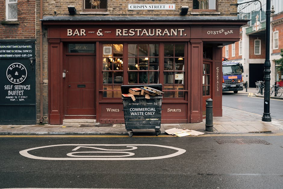 In front of a closed restaurant with a dark red wooden facade, there is a large black waste collection bin labeled 'Commercial Waste Only' positioned on the sidewalk. The bin contains cardboard boxes and miscellaneous rubbish, some of which are partially spilling onto the pavement. Surrounding the bin are a black bollard and a small post, both acting as street infrastructure. To the left, a blackboard sign advertises a self-service buffet with white lettering, mounted on the brick wall of an adjacent building. Behind the bin, the restaurant’s large glass windows reveal a warm interior with pendant lighting, and above, a street sign indicates 'Crispin Street.' The scene is set on a typical urban street with a narrow road marked by white lines and a large white motorway-style symbol painted on the asphalt in the foreground, indicating a no-stopping zone. A delivery truck and bicycles are visible in the background, emphasizing the urban context. Waste Removal Clapham’s service here relates to private disposal of commercial rubbish from hospitality establishments on high streets, supporting alternative waste handling outside typical municipal collection. The image depicts an on-street waste management scene in an urban setting, highlighting practices of private rubbish removal and collection for commercial premises.