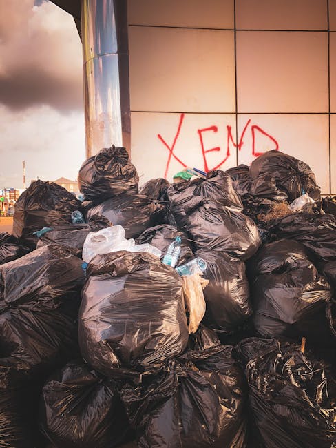 A large pile of black plastic rubbish bags is stacked against an exterior wall with a grid of beige tiles. The bags appear to be filled with various household waste and are piled irregularly, some leaning or resting on others. Several clear plastic water bottles are visible among the bags, some partially crushed or flattened, suggesting previous use and disposal. In the background, part of a metallic urban structure or pole reflects sunlight, and a cloudy sky is partially visible at the top left of the image. Red graffiti with the word 'XEND' is spray-painted on the tiled wall behind the rubbish, indicating an urban environment. The scene suggests an instance of private waste collection or on-site rubbish removal, potentially managed by Waste Removal Clapham, as evidenced by the informal nature of the waste containment and outdoor setting. The overall atmosphere is indicative of an area experiencing an accumulation of waste awaiting collection or disposal, aligning with rubbish removal services focused on bulk waste clearance.