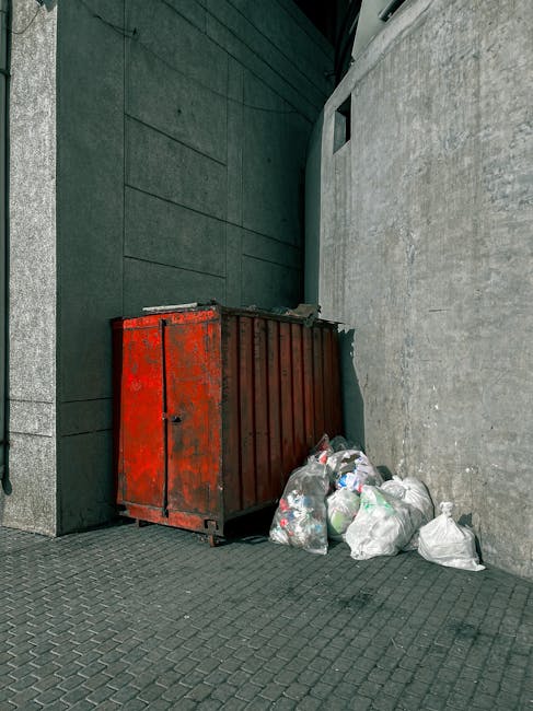 A red metal waste skip with a weathered surface showing patches of rust is positioned against a dark green wall on one side and a light grey concrete wall on the other, located on a paved surface with a textured pattern. The skip is partially filled with mixed rubbish and debris, while several plastic bags of waste, some appearing full and loosely tied, are piled beside it on the pavement. The environmental setting appears to be an urban alley or side street, with minimal natural light. The scene suggests an off-site, private waste collection or rubbish removal service provided by Waste Removal Clapham, designed for bulk or general waste disposal outside regular municipal collections, consistent with the context of alternative rubbish management options.