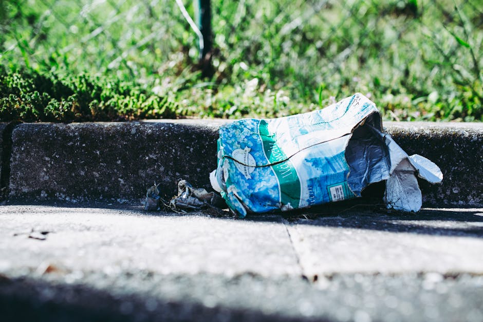 A crumpled plastic paper bag with a blue, white, and green design lies discarded on a concrete sidewalk near a dark curb. The bag appears faded and torn, with some of its contents visible through the damaged material. Small pieces of trash are scattered around it. Behind the curb, there is a patch of green grass with some blurred leafy plants, indicating an outdoor setting. The lighting suggests natural daylight, casting soft shadows on the pavement. This scene illustrates the presence of improperly disposed waste in a public or residential outdoor area, highlighting the need for waste removal services like those offered by Waste Removal Clapham, which may involve private or alternative waste handling methods for rubbish clearance.
